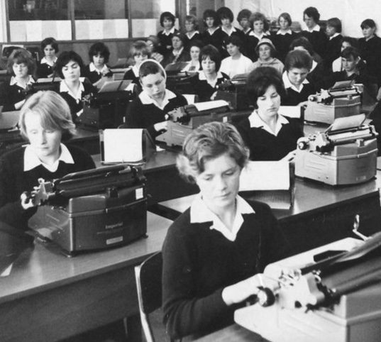 Black and white photo of teenage students sat at typewriters in a classroom while other students are standing in the background